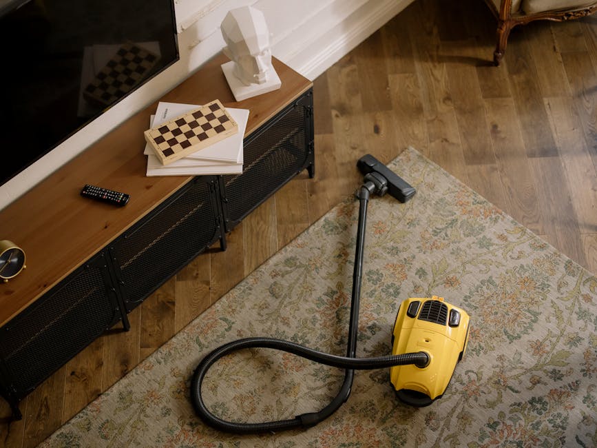 A child using a lightweight, handheld vacuum cleaner to perform surface cleaning on a patterned, traditional-style carpet in a residential living room. The carpet features a floral design in muted ton