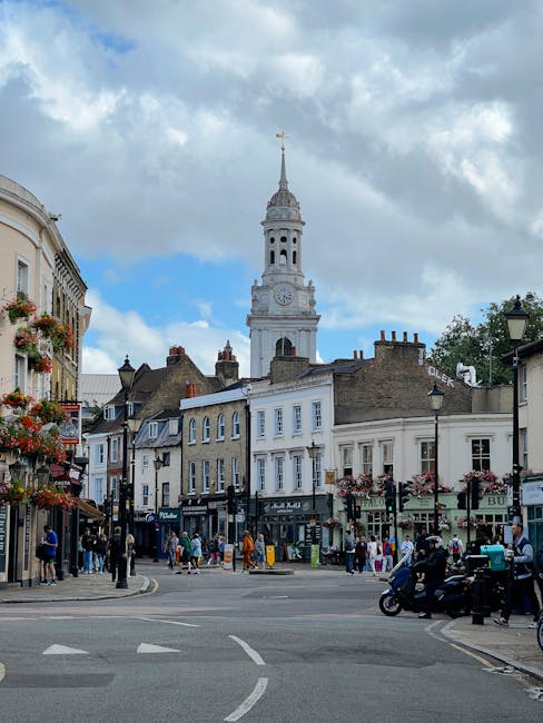 A busy street scene in North Cross Road, East Dulwich, featuring a mix of small shops and cafes with storefronts decorated with hanging flower baskets. In the background, a tall white clock tower with